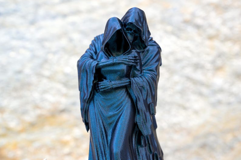 May include: A dark blue, monochromatic figurine depicting two hooded, skeletal figures in a close embrace. The figures are draped in long, flowing robes with visible folds, and the skeletal hands of one figure are wrapped around the other's torso.