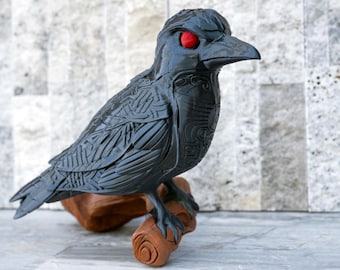May include: A dark gray raven sculpture with intricate carved details and bright red eyes. The bird is perched on a brown, wooden-looking base. The sculpture has a detailed feather pattern and is a decorative item.