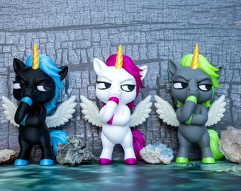 Puede incluir: Tres figuras de unicornios con alas, cada una con un cuerno dorado y una mano cubriendo su boca. La figura de la izquierda es negra con pelo azul, la central es blanca con pelo rosa y la de la derecha es gris con pelo verde lima.