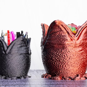 May include: Two decorative pen holders shaped like alien eggs. One is black and filled with pens and markers. The other is a reddish-brown color and filled with colorful candies. Both have a textured surface and a unique design.