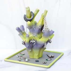 May include: A coral-shaped jewelry stand with a gradient color scheme transitioning from lime green at the base to light purple at the tips. The stand features multiple branches for hanging necklaces and earrings, set on a square tray base.