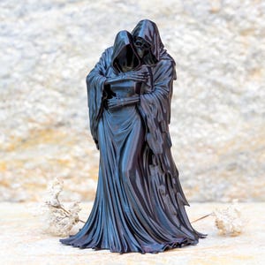 May include: A black figurine depicting two hooded figures in a close embrace. The figures are draped in long, flowing robes that reach the ground, with skeletal hands visible as they hold each other. The sculpture has a smooth, dark finish.