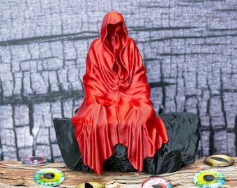 Puede incluir: Una escultura de figura con capucha roja se asienta sobre una base de roca negra. La figura está envuelta en una capa roja larga y fluida que cubre todo el cuerpo. Varios ojos de cabujón de vidrio con diversos colores de iris están esparcidos.