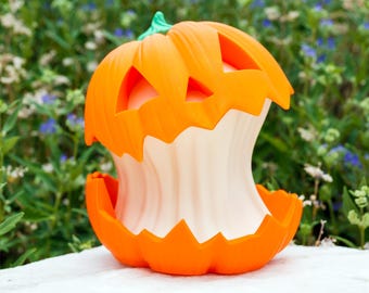 Puede incluir: Una decoración de calabaza jack-o'-lantern de color naranja brillante con un tallo verde, que presenta un diseño dentado y abierto que revela un núcleo interior liso de color crema. La calabaza tiene ojos triangulares y una boca ancha.