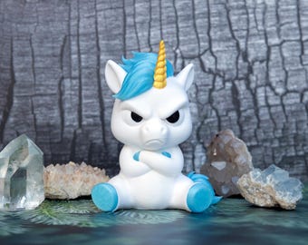 Puede incluir: Una figura de unicornio blanco con crin azul brillante, cuerno en espiral amarillo y cascos azules. El unicornio tiene una expresión facial severa con cejas fruncidas y brazos cruzados sobre el pecho, sentado entre varios grupos de cristales.