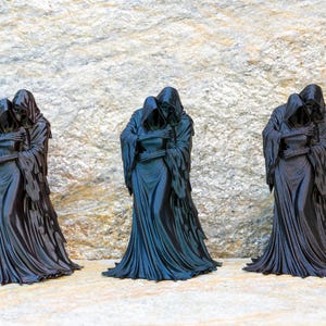 May include: Three identical black figurines depicting two hooded figures in a close embrace. Each sculpture features long, flowing robes with detailed folds and textures, capturing a somber and unified stance of two cloaked entities standing together.