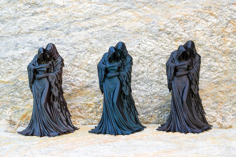 May include: Three identical black figurines depicting two hooded figures in a close embrace. Each sculpture features long, flowing robes with detailed folds and textures, capturing a somber and unified stance of two cloaked entities standing together.