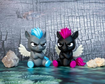 Puede incluir: Dos pequeñas figuras de poni alado con expresión gruñona sentadas una al lado de la otra. El poni de la izquierda es gris con crines, cola y cascos azul brillante. El de la derecha es negro con crines, cola y cascos color magenta.