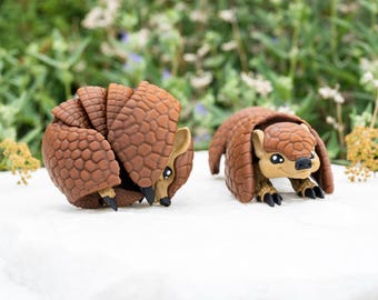 Puede incluir: Dos figuras de armadillo de color marrón con caparazones segmentados y texturizados. Una figura está enrollada en una bola apretada, mientras que la otra está en posición de pie con la cabeza y las garras delanteras visibles, mostrando escamas.