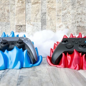 May include: Two game controllers, one silver and one black, each resting on a stylized flame-shaped base. The silver controller sits on a blue base, while the black controller is on a red base. The background is a stone wall.