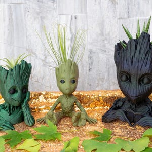 May include: Three Groot-inspired planters in green and black, each holding a different plant. The planters have detailed wooden textures and expressive faces. The plants include air plants and a succulent. The planters are on a gold sequined surface.
