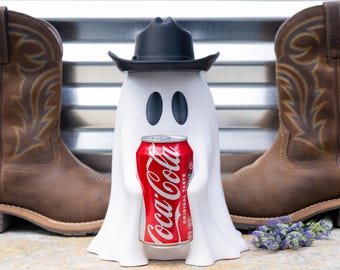 Puede incluir: Una figura de fantasma blanca con un sombrero de vaquero negro que sostiene una lata roja de Coca-Cola, colocada entre dos botas de vaquero de cuero marrón. El fantasma tiene dos ojos ovalados negros y un acabado mate liso, de pie en el centro.