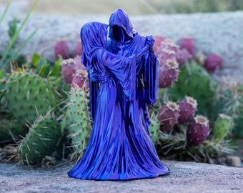 Puede incluir: Una figura de color azul metálico que representa a dos personajes encapuchados en un abrazo danzante. Las figuras visten largas túnicas fluidas con pliegues profundos y sus manos esqueléticas son visibles mientras se sostienen en posición erguida.