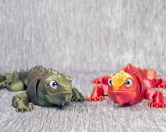 May include: Two toy lizards, one green and one red, are displayed on a textured gray surface. The green lizard has purple eyes, while the red lizard has blue eyes and an orange head. Both are stylized.