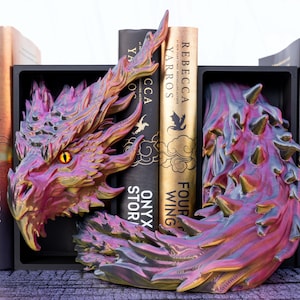 May include: A fantasy-themed bookend set featuring a detailed dragon design in shades of purple, pink, and gold. The dragon's head and tail frame the books, with titles like "Iron Flame" and "Fourth Wing" visible. The bookends are black.