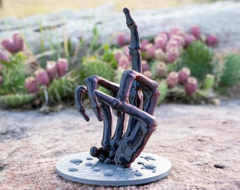 Puede incluir: Una escultura de mano esquelética de color morado oscuro y brillante con el dedo corazón extendido en un gesto vertical. La mano está unida a una base circular de color gris claro con pequeñas texturas en relieve similares a guijarros.