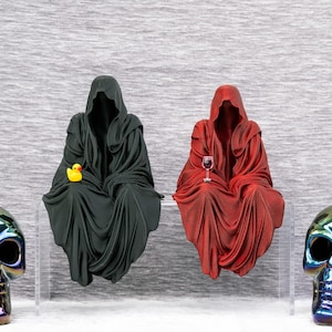 May include: A whimsical composition featuring two hooded figures, one black and one red, seated with a rubber duck and a wine glass. Flanking them are iridescent skull sculptures, creating a unique, gothic-inspired aesthetic.