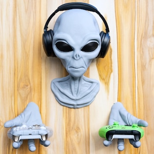 May include: A gray alien head with black headphones and two gray hands holding video game controllers. One controller is gray, the other is green. The alien is mounted on a wooden surface.