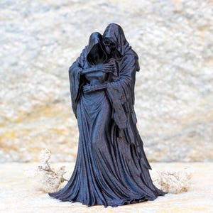 May include: A black figurine depicting two hooded skeletal figures in a close embrace. The figures are draped in long, flowing robes with intricate folds and textures. The taller figure stands behind the other, with skeletal hands resting on the shoulders.