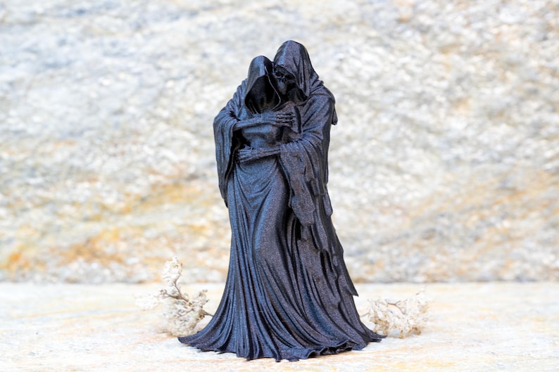 May include: A black figurine depicting two hooded skeletal figures in a close embrace. The figures are draped in long, flowing robes with intricate folds and textures. The taller figure stands behind the other, with skeletal hands resting on the shoulders.