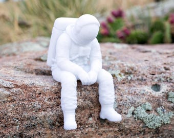 Puede incluir: Una figura blanca mate de un astronauta en posición sentada con la cabeza inclinada, vistiendo un traje espacial detallado con mochila de soporte vital, casco y botas texturizadas, creada como un objeto decorativo sólido.
