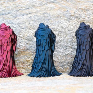 May include: Three standing figurines depicting hooded figures in long, layered cloaks. The left figurine is metallic magenta, the center is metallic blue, and the right is matte black. Each figure features a draped, textured garment design.