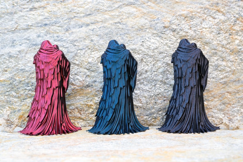 May include: Three standing figurines depicting hooded figures in long, layered cloaks. The left figurine is metallic magenta, the center is metallic blue, and the right is matte black. Each figure features a draped, textured garment design.
