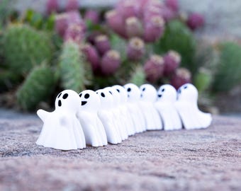 Puede incluir: Una fila de nueve pequeñas figuras de fantasmas blancos dispuestas en línea. Cada fantasma presenta una cabeza lisa y redondeada con dos ojos ovalados negros y una boca ovalada negra. Las figuras están sobre una superficie de piedra texturizada.