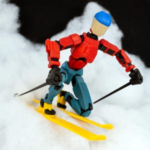 May include: A red, blue, and black articulated action figure wearing a blue helmet is skiing down a snowy slope. The figure is holding two ski poles and is in a crouched position.