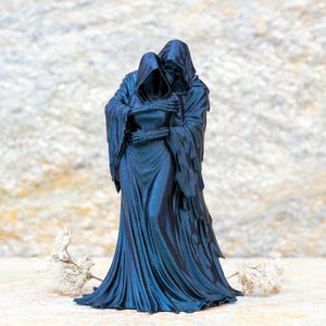 May include: A dark blue, monochromatic figurine depicting two hooded, skeletal figures in a close embrace. The figures are draped in long, flowing robes with intricate folds, and their bony hands are visible as they hold each other tightly.
