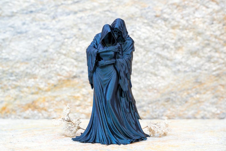 May include: A dark blue, monochromatic figurine depicting two hooded, skeletal figures in a close embrace. The figures are draped in long, flowing robes with intricate folds, and their bony hands are visible as they hold each other tightly.