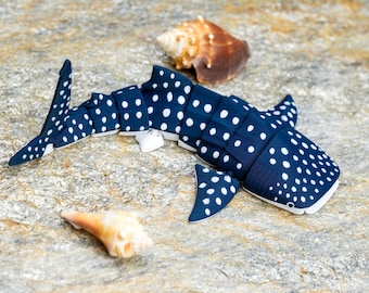 Puede incluir: Una figura articulada de tiburón ballena azul oscuro con un patrón de puntos blancos en el cuerpo, las aletas y la cola. El tiburón está colocado cerca de dos conchas marinas de color tostado y marrón, con un diseño segmentado y flexible.