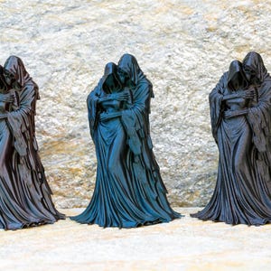 May include: Three identical black figurines depicting two hooded figures in a close embrace. Each sculpture features draped, flowing robes with intricate folds and skeletal hands resting on the shoulders of the other figure, standing upright.