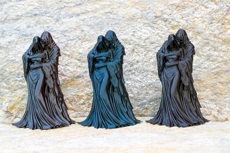 May include: Three identical black figurines depicting two hooded figures in a close embrace. Each sculpture features draped, flowing robes with intricate folds and skeletal hands resting on the shoulders of the other figure, standing upright.
