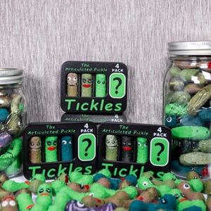 May include: A display of novelty items featuring green, blue, and brown articulated pickle figures. Some are in jars, others are loose, and some are packaged in black boxes with the word "Tickles" and a question mark.