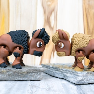 May include: Two brown bison figurines with black and tan fur details, standing on gray stone platforms. The bison have large eyes and are posed facing each other. The background is a light wood grain.