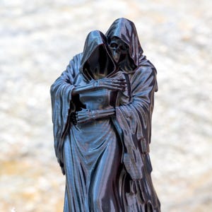 May include: A glossy black figurine depicting two hooded skeletal figures in a close embrace. The figures are draped in long, flowing robes with visible folds and textures, and their bony hands are clasped together against the chest of the other.