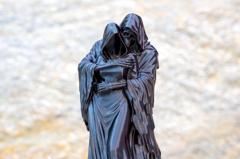May include: A glossy black figurine depicting two hooded skeletal figures in a close embrace. The figures are draped in long, flowing robes with visible folds and textures, and their bony hands are clasped together against the chest of the other.