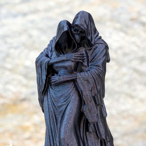 May include: A black, glittery figurine depicting two hooded skeletal figures in a close embrace. The figures are draped in long, flowing robes with visible skeletal hands resting on each other's shoulders and backs, creating a somber composition.