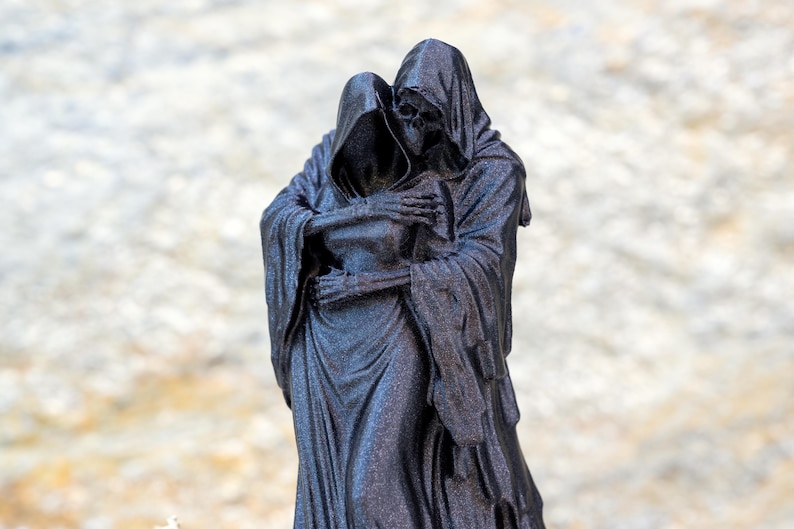 May include: A black, glittery figurine depicting two hooded skeletal figures in a close embrace. The figures are draped in long, flowing robes with visible skeletal hands resting on each other's shoulders and backs, creating a somber composition.