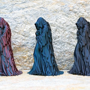 May include: Three standing figurines of hooded figures in long, draped robes. The left figure is a metallic reddish-purple, the center figure is a metallic blue, and the right figure is a solid matte black. Each figure has a draped hood.