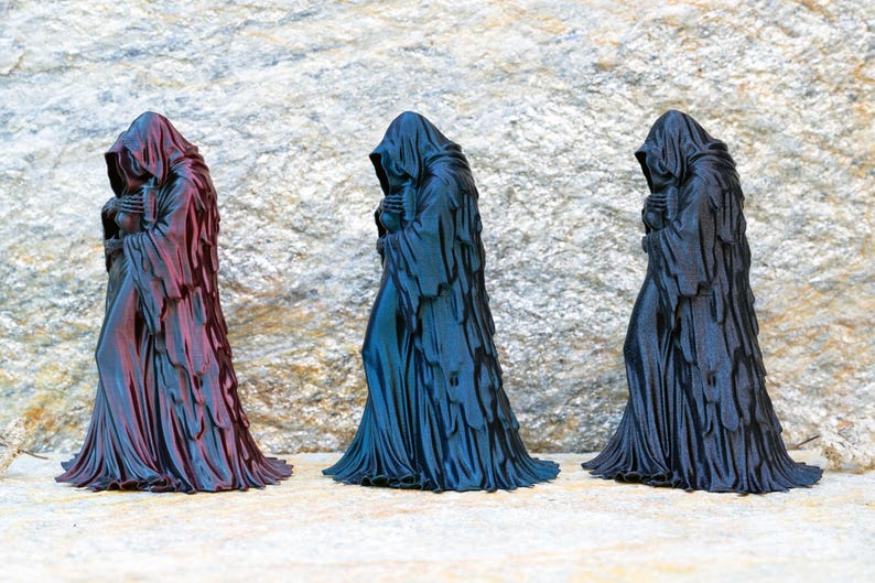 May include: Three standing figurines of hooded figures in long, draped robes. The left figure is a metallic reddish-purple, the center figure is a metallic blue, and the right figure is a solid matte black. Each figure has a draped hood.
