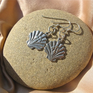 May include: A pair of silver seashell earrings. Each earring features a detailed, fan-shaped shell design with a small floral accent. The earrings are suspended from silver hooks and are displayed on a smooth, light brown stone, set against a backdrop of a peach-colored fabric.