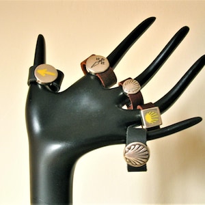 May include: Five leather bracelets with silver metal charms. The charms feature a yellow arrow, a yellow sun, and a scallop shell.