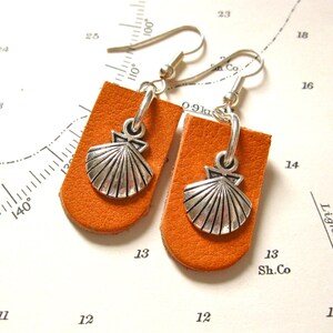 May include: A pair of orange leather earrings with silver seashell charms. The earrings have a hook closure.