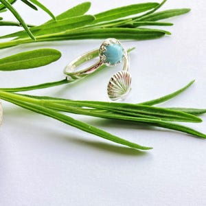 May include: A silver ring with a light blue gemstone and a shell design. The ring is on a white background with green foliage.