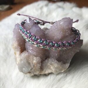 May include: A multi-coloured friendship bracelet with white, purple and teal threads woven together in a knot pattern. The bracelet is displayed on a cluster of amethyst crystals.
