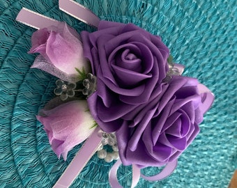 May include: A purple and white artificial flower wrist corsage with a satin ribbon and pearl accents.