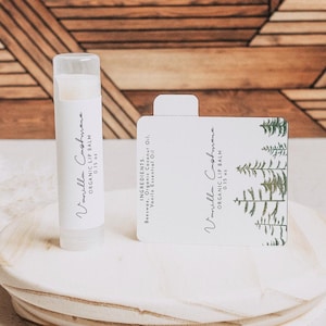 Puede incluir: Un tubo blanco de bálsamo labial orgánico Vanilla Cashmere con una etiqueta verde y blanca que representa una escena de bosque. El bálsamo labial está sobre una plataforma de madera.