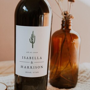 以下が含まれることがあります： 緑色のサボテンのイラストが描かれたカスタムラベルのワインボトル。ラベルには「08.07.20XX Isabella & Harrison Moab, Utah」と記載されています。